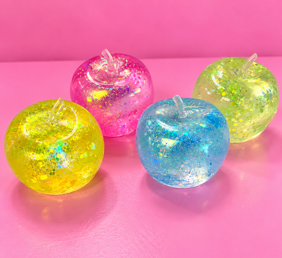 Four colourful clear glitter apple squishies on a pink background